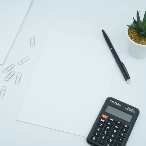 A picture of a black wooden table with a black pocket calculator, 2 white sheets of paper and a small white cactus pot