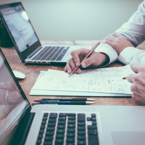 A picture of 2 people at a table with 2 computers and a picture of accounting graphs on them, you can only see hands, one hand with a pencil towards a page with some text